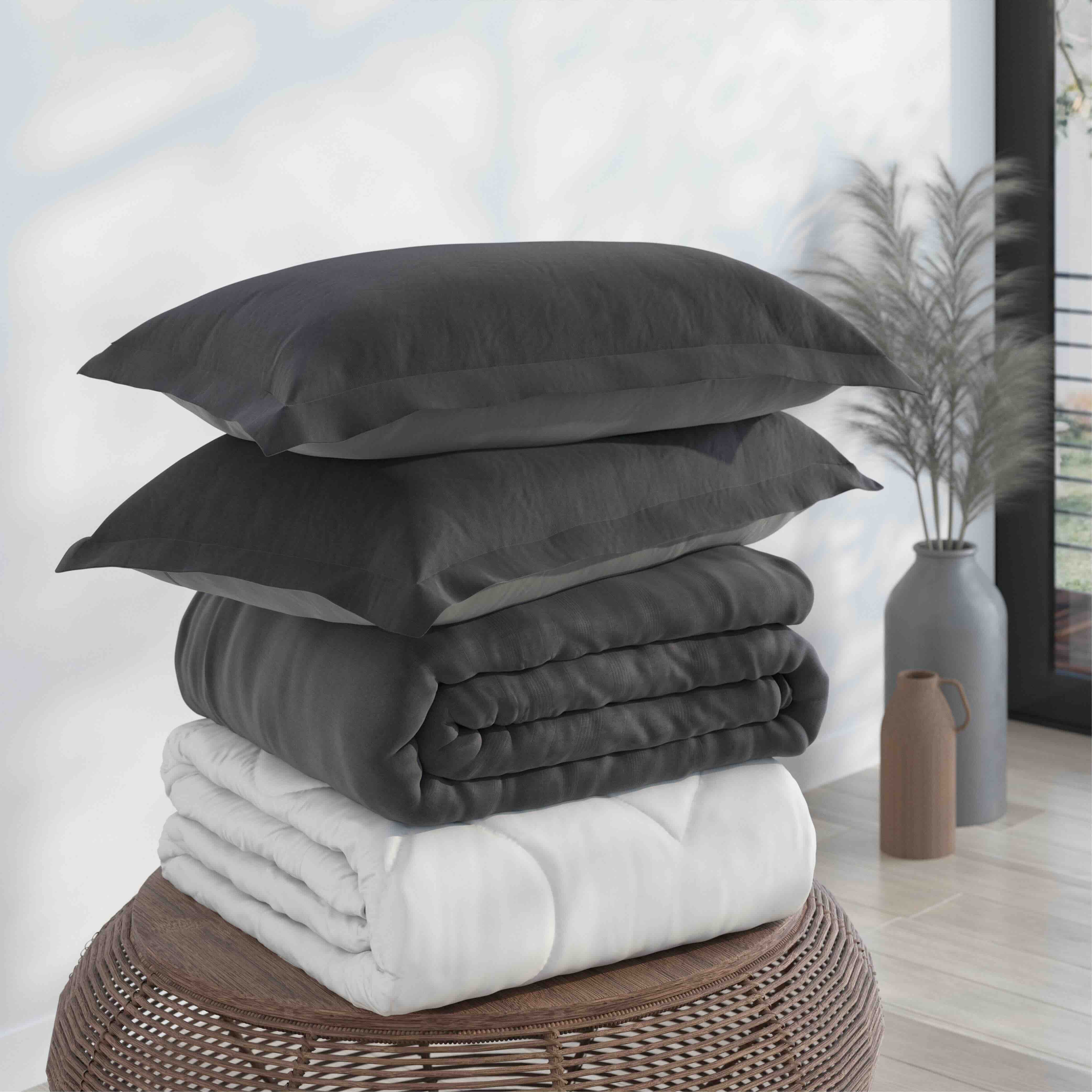 A stack of dark gray pillows and folded bedding sits on a woven round stool in a bright room, with a white folded blanket underneath and a vase of dried stems in the background.