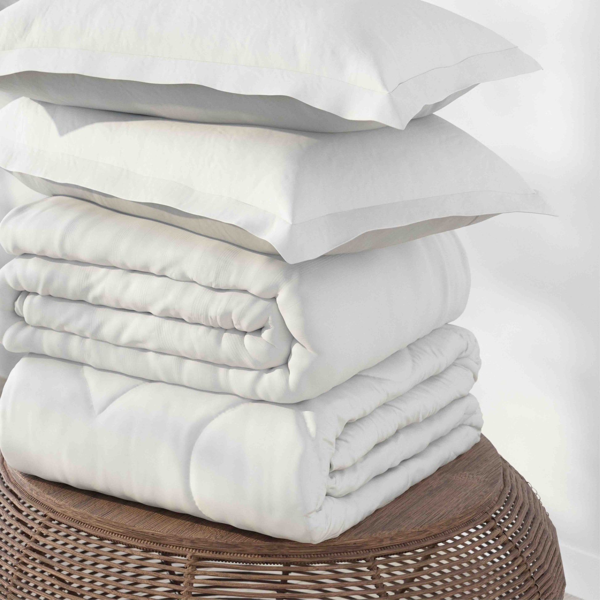 A stack of white bedding, including two pillowcases and duvet cover, displayed on a round woven wicker stool against a plain white background.