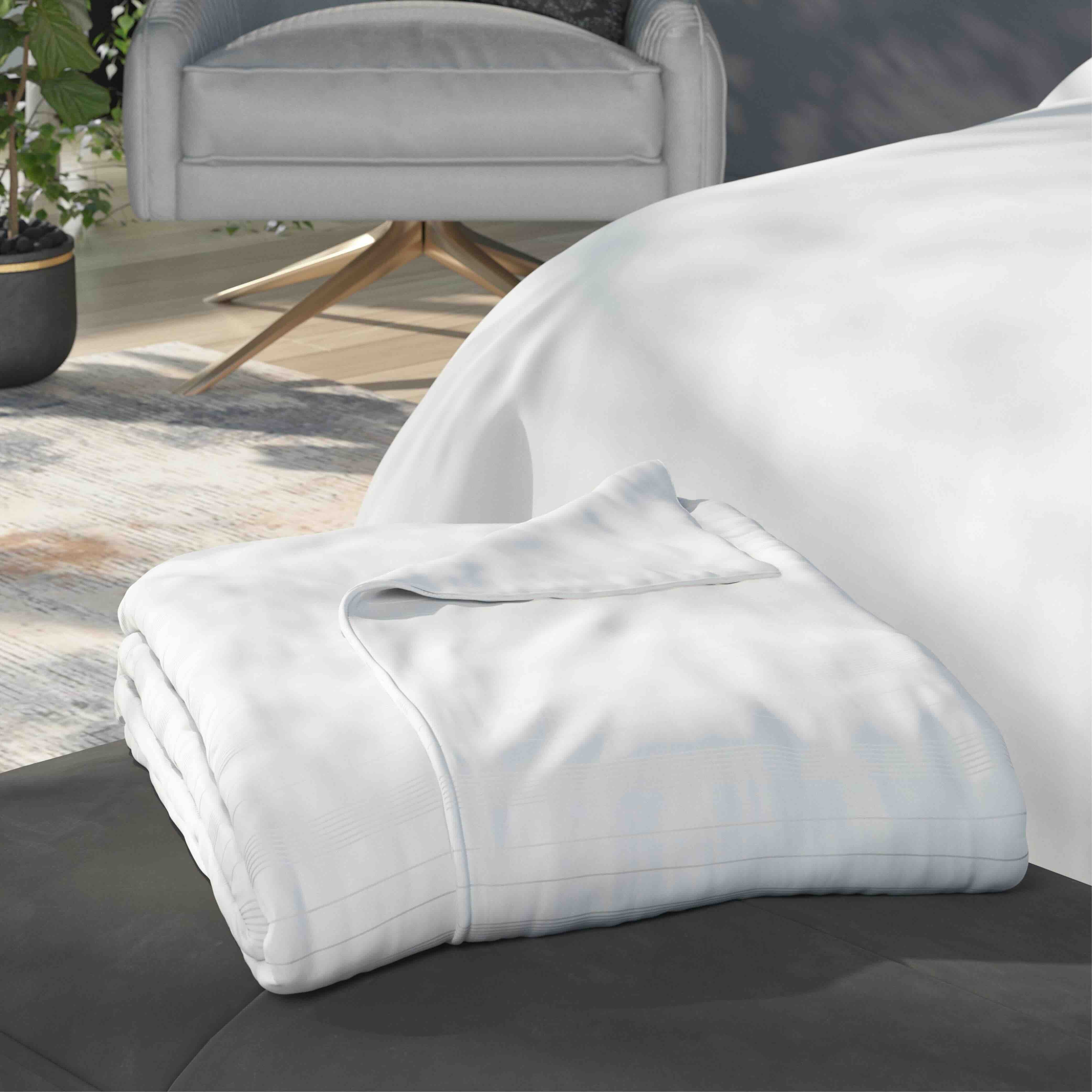 A folded white duvet cover sits on a dark upholstered bench in a modern bedroom setting, with a bed covered in white bedding and a gray armchair in the background.