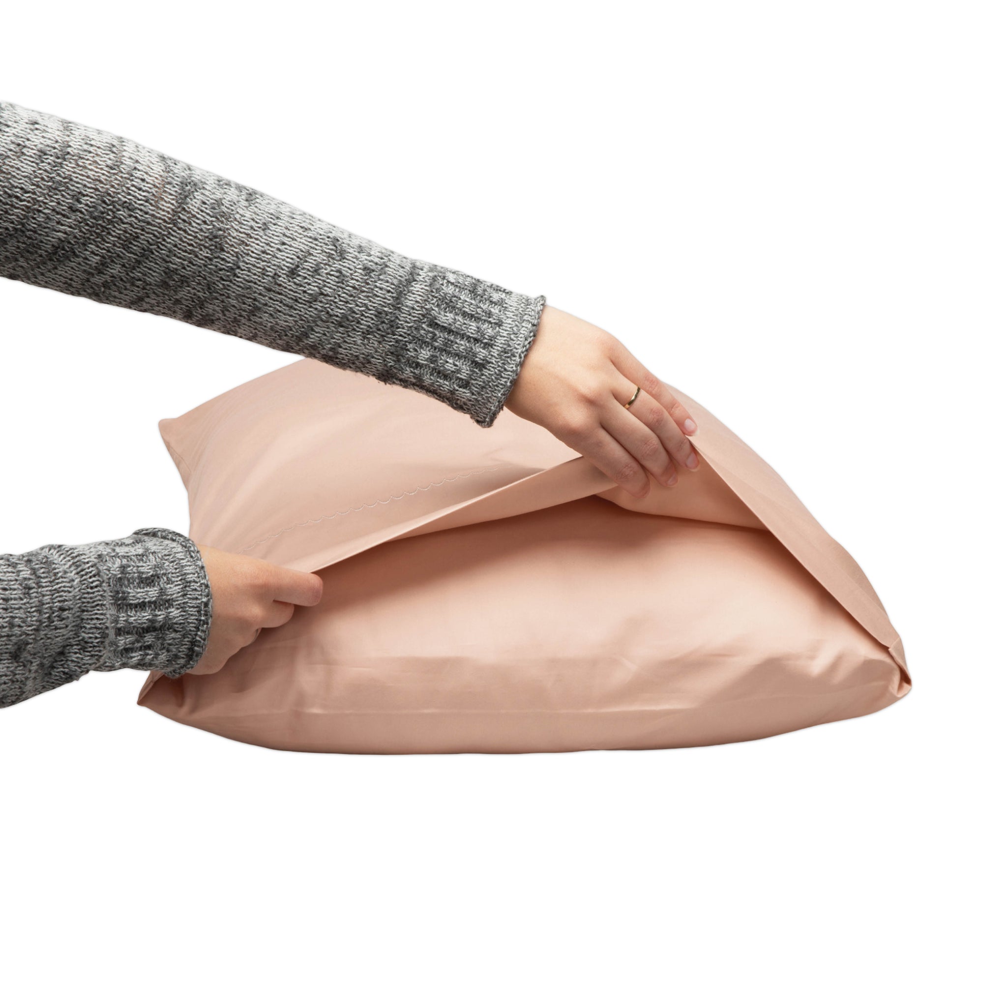 A person wearing a gray knit sweater is shown from the shoulders down, opening a dusty pink pillowcase on a white background.