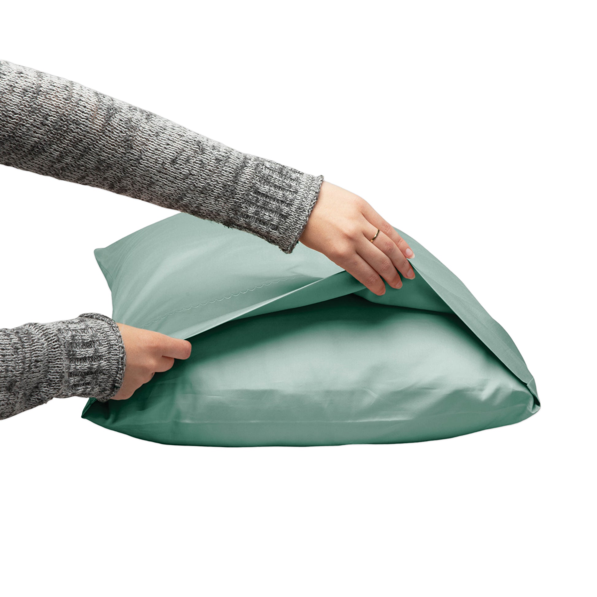 A person wearing a gray knit sweater is opening a sage green pillowcase on a white background.