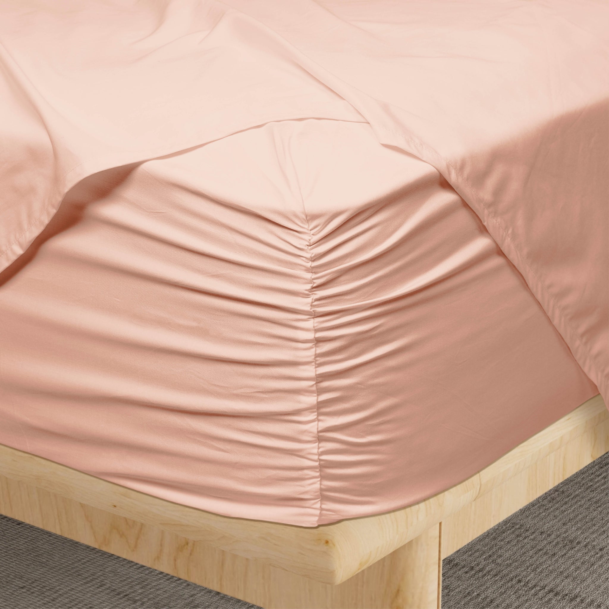 A close-up of a blush pink fitted sheet tucked tightly around a wooden bed frame, showing gathered fabric at the corner and a gathered pocket seam.