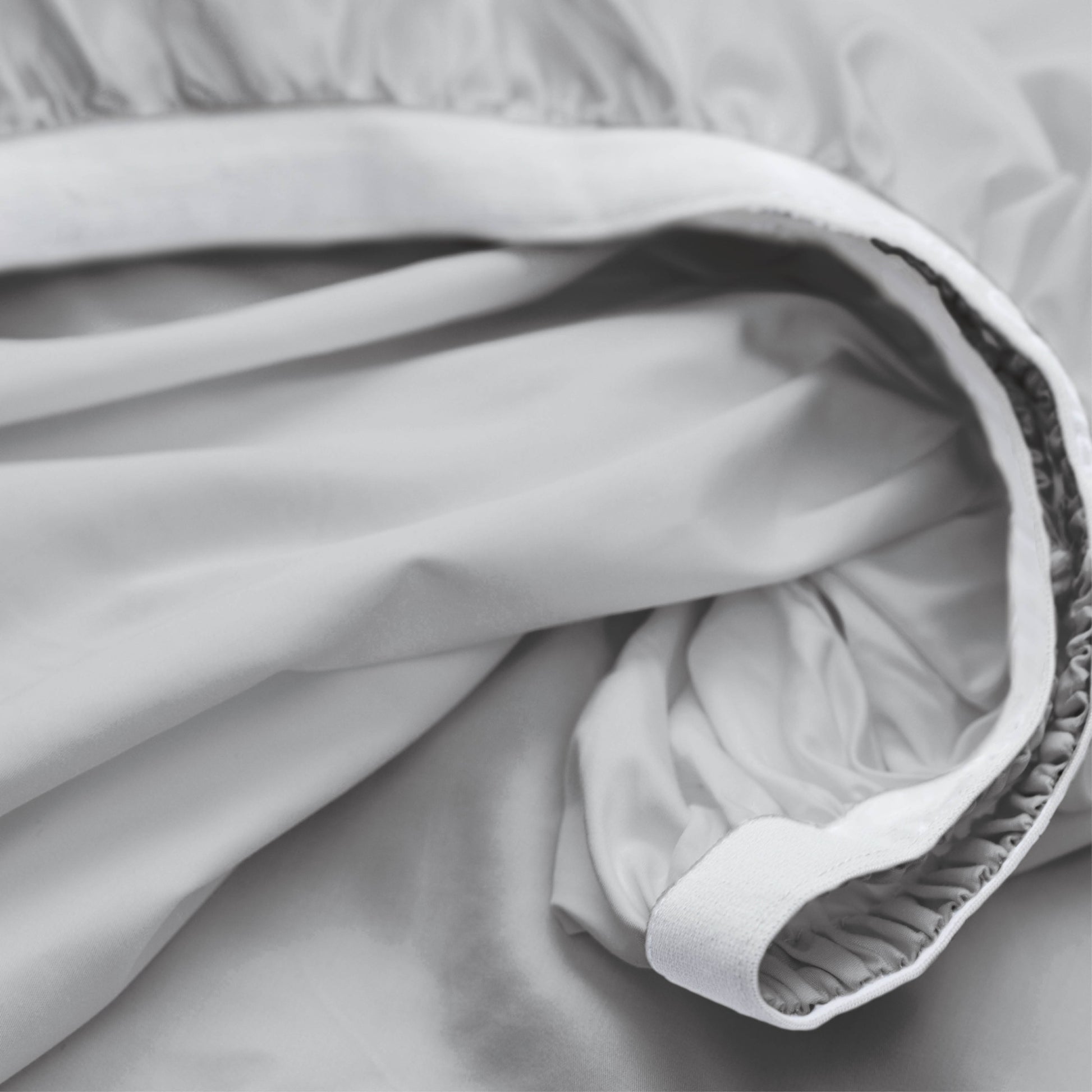 Close-up of a light gray or white bedding fabric, likely a duvet cover or sheet, showing the gathered elasticized edge and smooth draped texture.