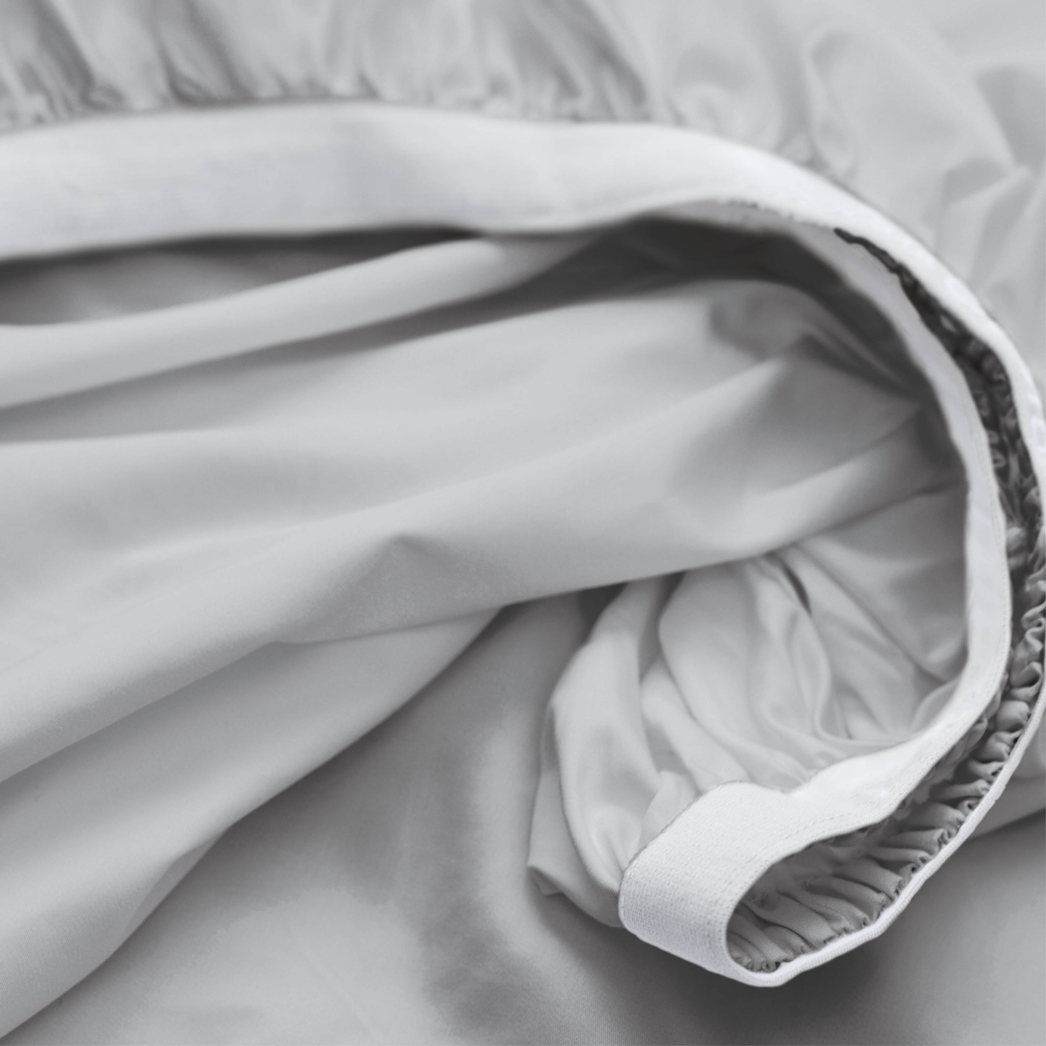 Close-up of a light gray or white bedding fabric, likely a duvet cover or sheet, showing the gathered elasticized edge and smooth draped texture.