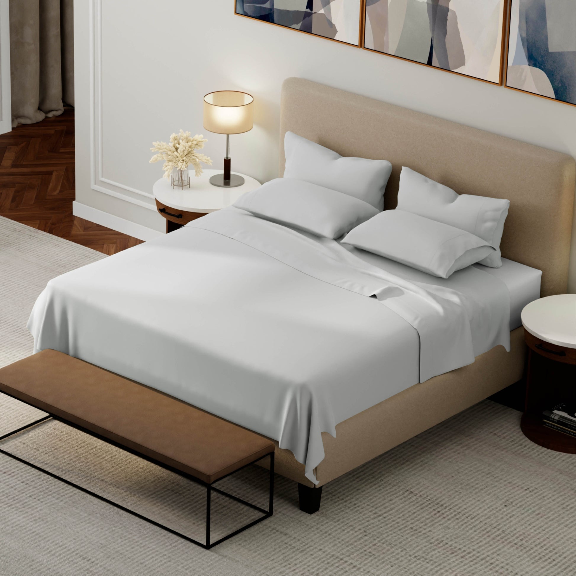 A neatly made bed with grey bedding and pillows, set in a modern bedroom with a beige upholstered headboard, bedside tables, a lamp, and a bench at the foot of the bed.