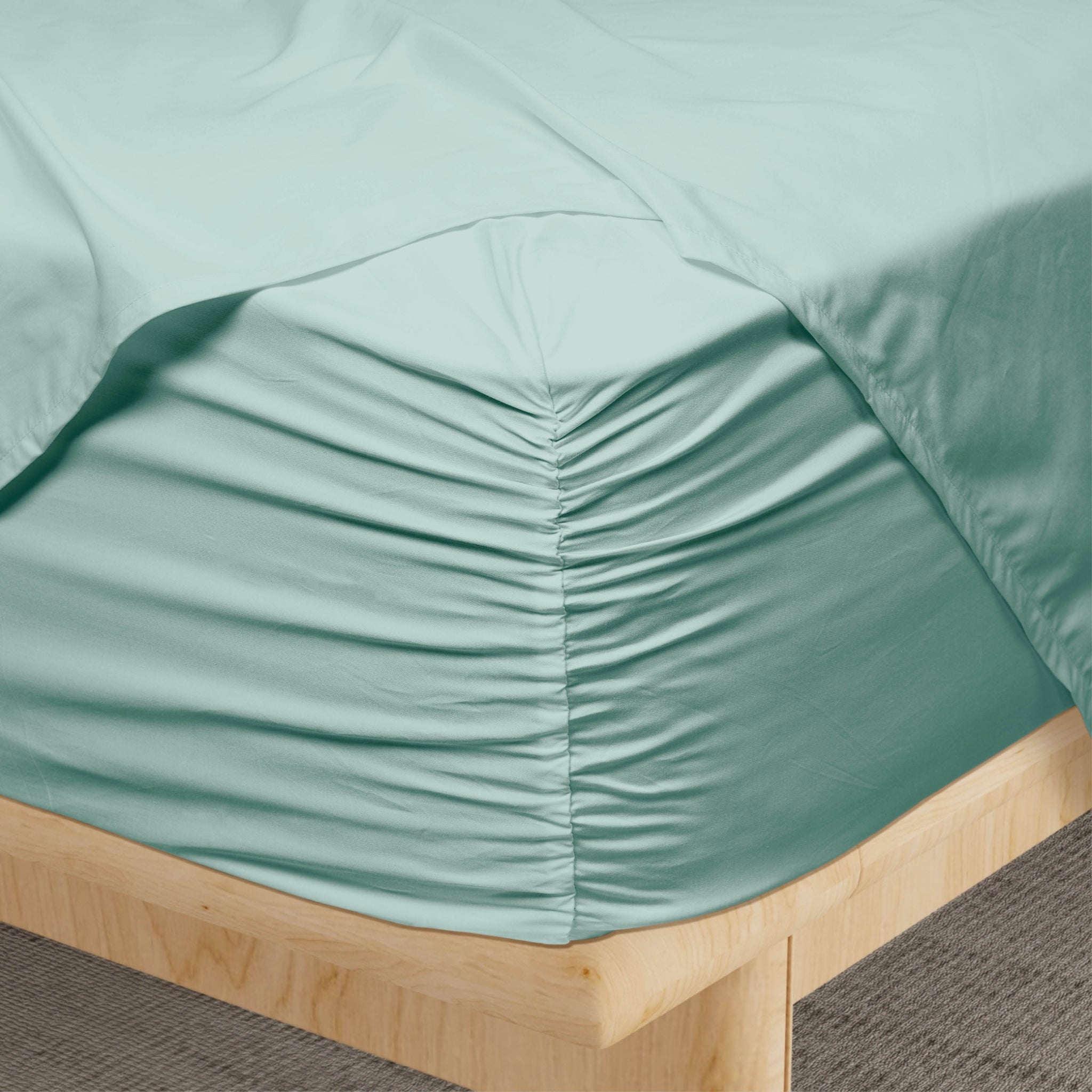 A close-up of a fitted sheet in a muted sage green color, gathered and ruched around the corner of a bed with a light wood frame.