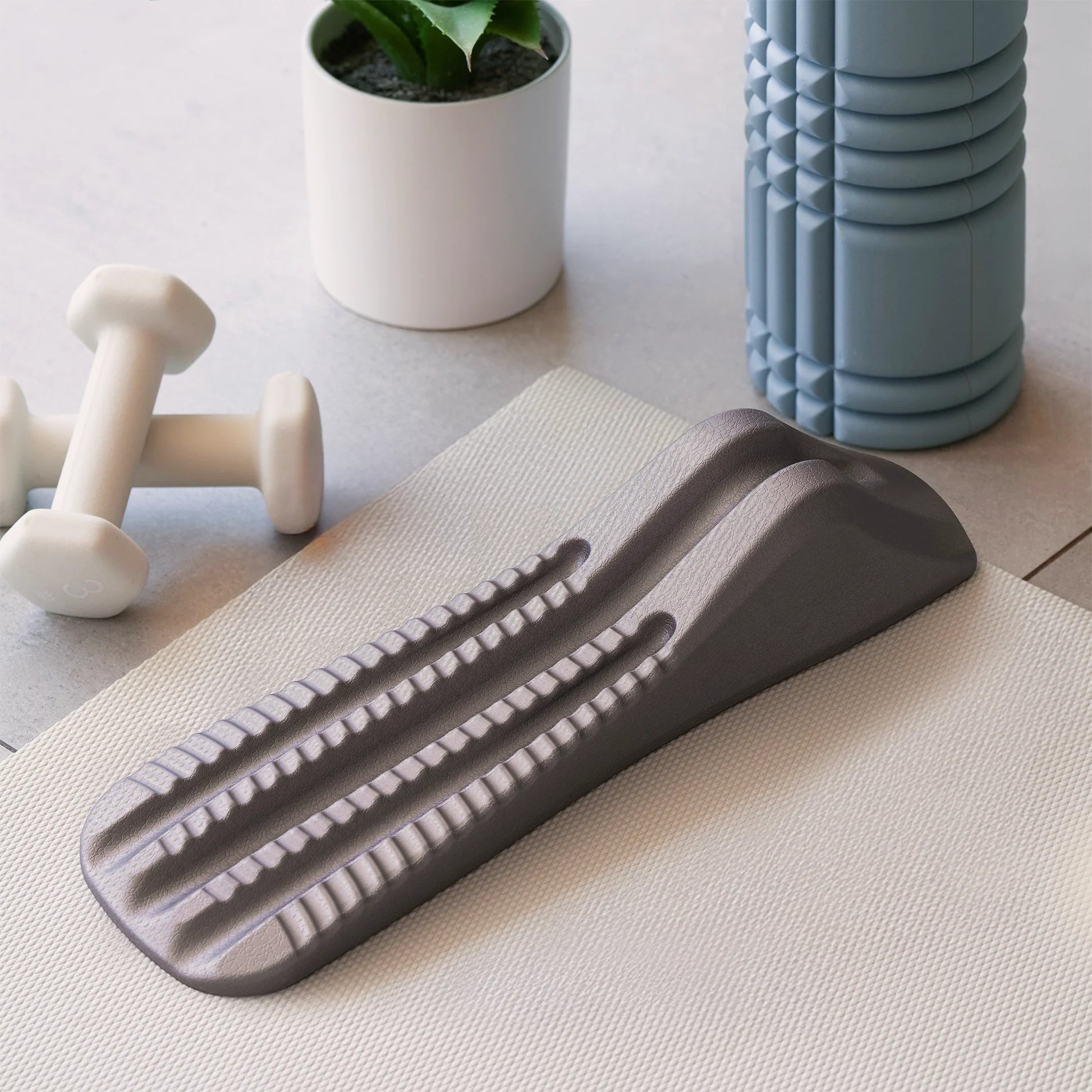 Gray spine decompression stretcher on a light surface with dumbbells and a plant in the background.