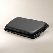 A black rectangular padded footrest with a smooth textured top surface and a slightly raised base, photographed on a plain light beige studio background.
