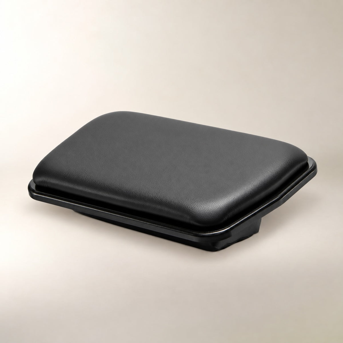 A black rectangular padded footrest with a smooth textured top surface and a slightly raised base, photographed on a plain light beige studio background.