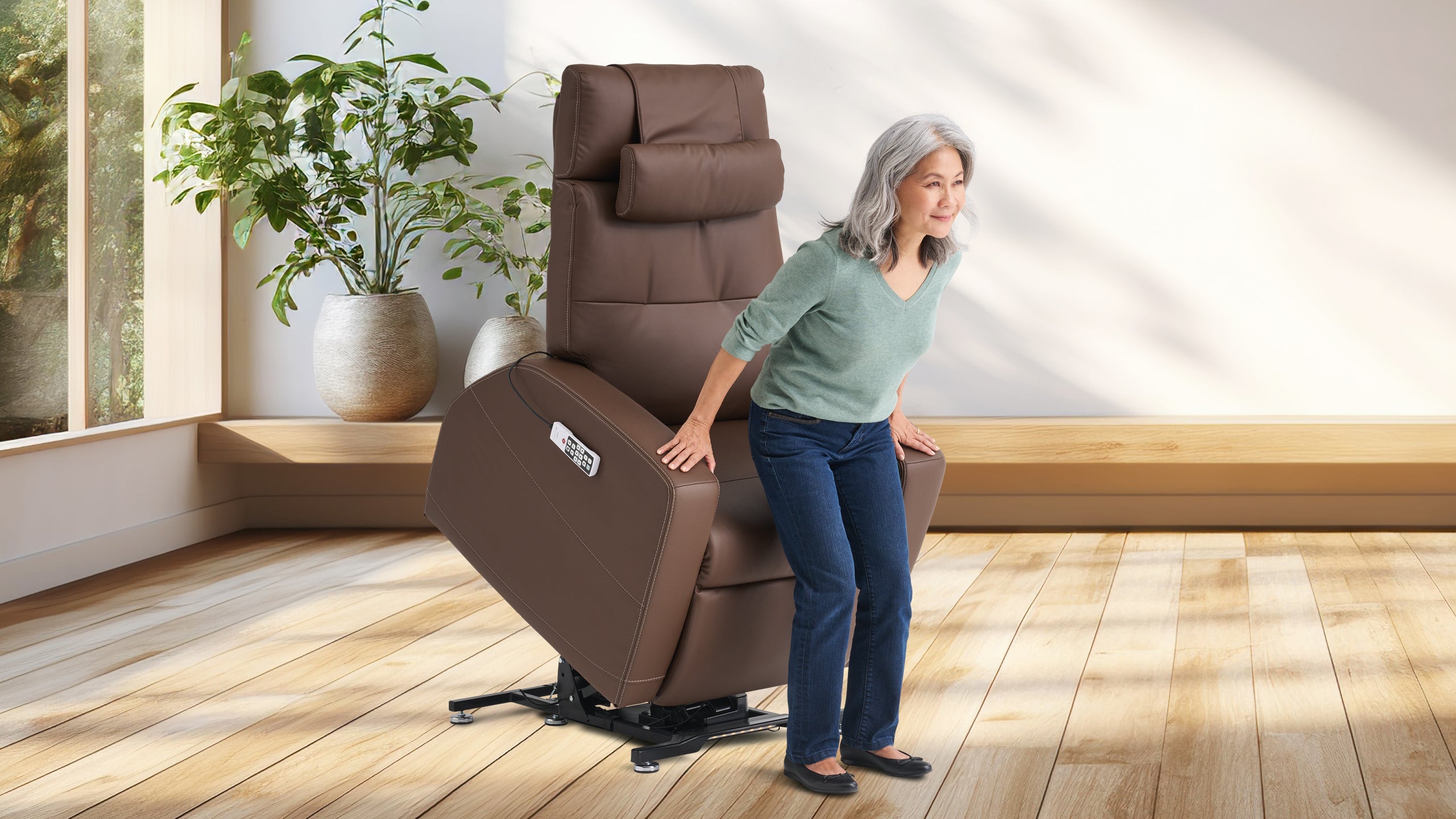 Older woman using a brown power lift recliner chair in a bright room with wood flooring and large potted plants. The chair is tilted forward in lift position to assist her in standing up, with a wired remote attached to the side.
