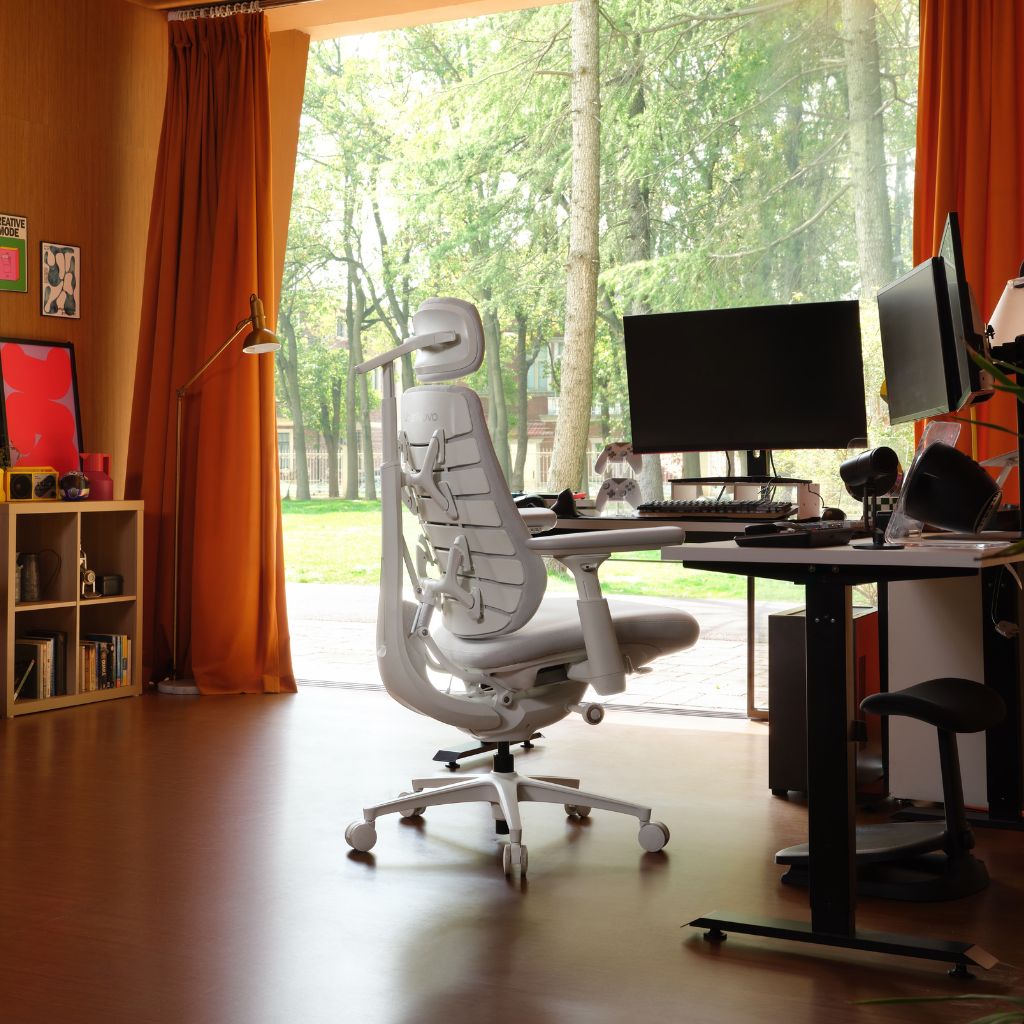 A white ergonomic office chair with a headrest and lumbar support is positioned in front of a black adjustable desk in a home office setup. The room features warm orange walls, orange curtains, and a large window looking out onto a green wooded area.
