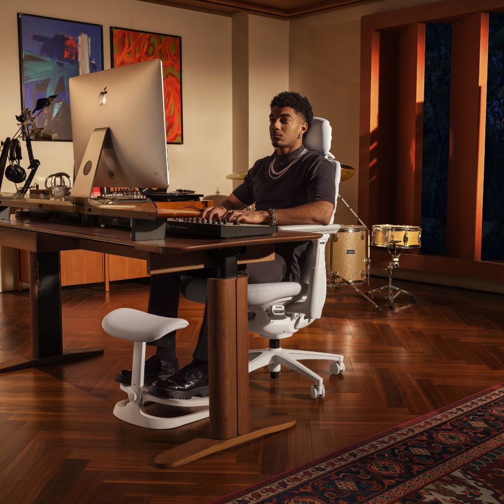 A man sits in a white ergonomic office chair at a wooden desk in a warm, modern home studio. He is using a computer and has a footrest under his foot.