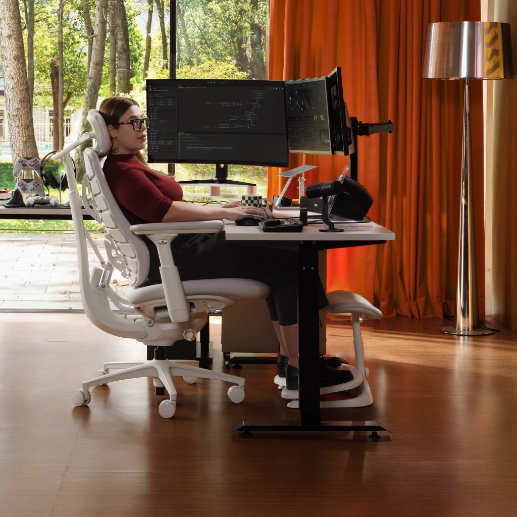 A woman sits in a white ergonomic office chair at a standing desk with dual computer monitors, in a modern room with orange curtains, a floor lamp, and a large window looking out onto trees.