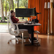 A woman sits in a white ergonomic office chair at a standing desk with dual computer monitors, in a modern room with orange curtains, a floor lamp, and a large window looking out onto trees.
