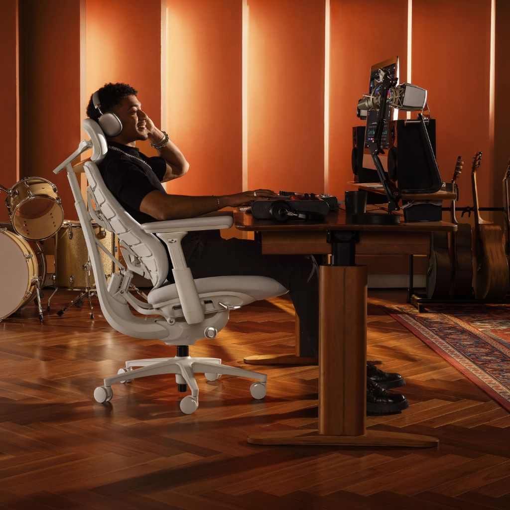 A man sits in a white ergonomic office chair at a wooden desk with dual computer monitors, in a warm, orange-lit home office with musical instruments in the background.