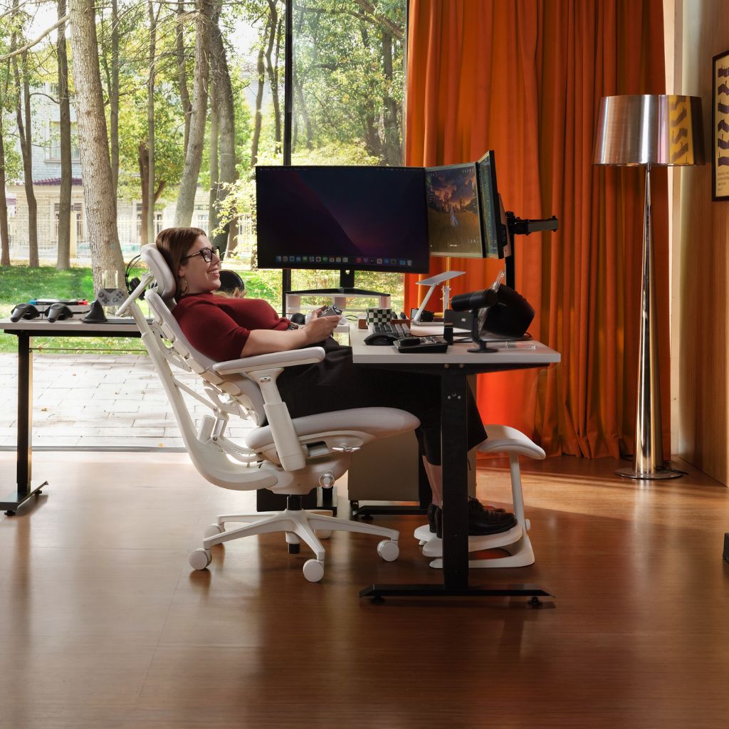 A woman sits in a white ergonomic office chair at a height-adjustable standing desk with dual computer monitors, in a bright room with large windows, orange curtains, and a floor lamp.