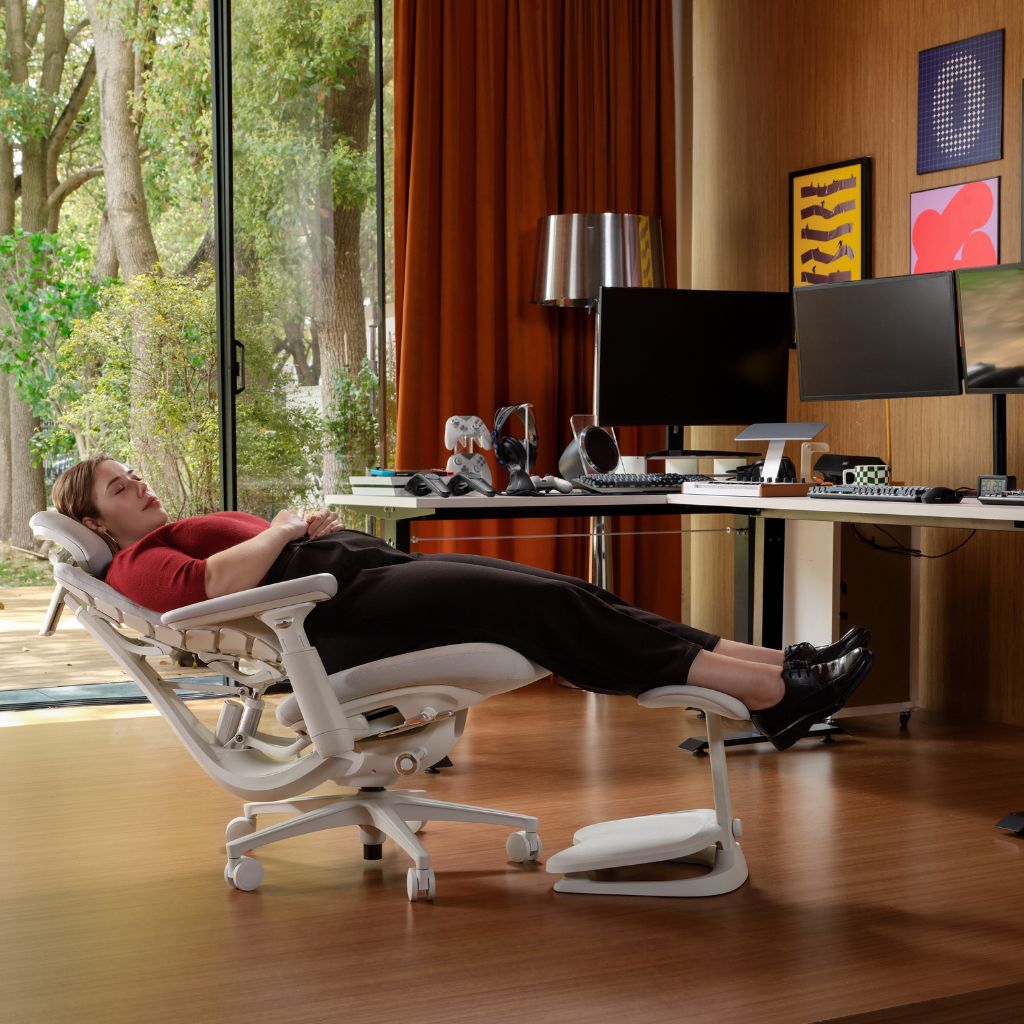 A woman reclines in a white ergonomic office chair with extended footrest and headrest, set in a modern home office with wood floors, desk, computer monitors, and large windows.