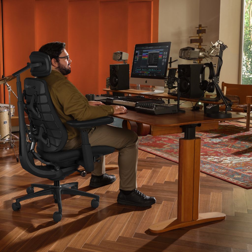 A man sits in a black ergonomic office chair at a wooden desk in a home studio setup, with a computer monitor, speakers, and recording equipment visible in a room with orange walls and wood flooring.