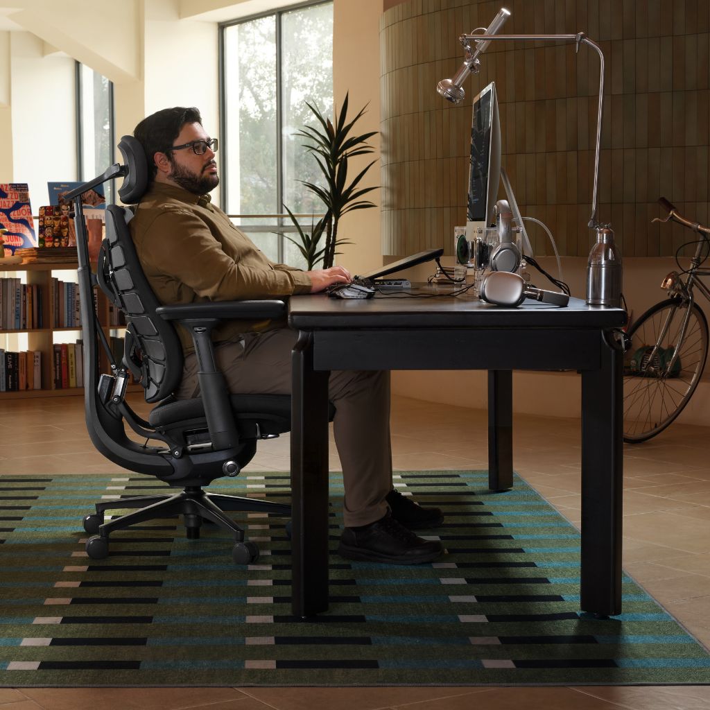A man sits in a black ergonomic office chair at a dark desk in a modern home office, using a computer. The room features a large patterned green and black rug, a potted plant, and a bicycle against a tiled wall.