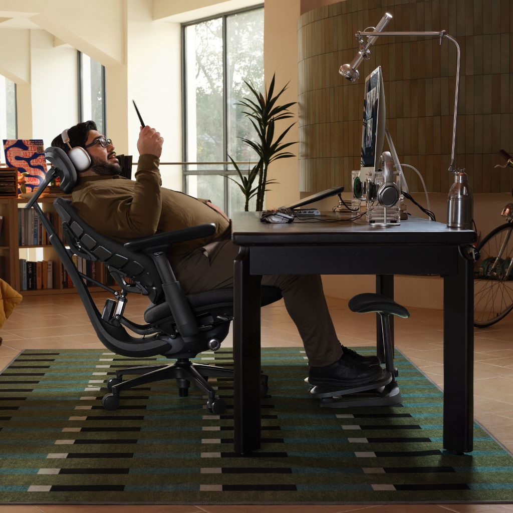 A man reclining in a black ergonomic office chair at a dark desk with a computer monitor, headphones, and other office accessories in a modern home office setting.