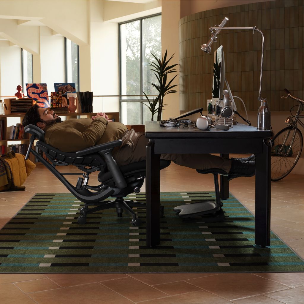A man reclines in a black ergonomic office chair at a dark desk in a modern home office setting. The room features a green and black patterned rug, large windows, and a bicycle leaning against the wall.