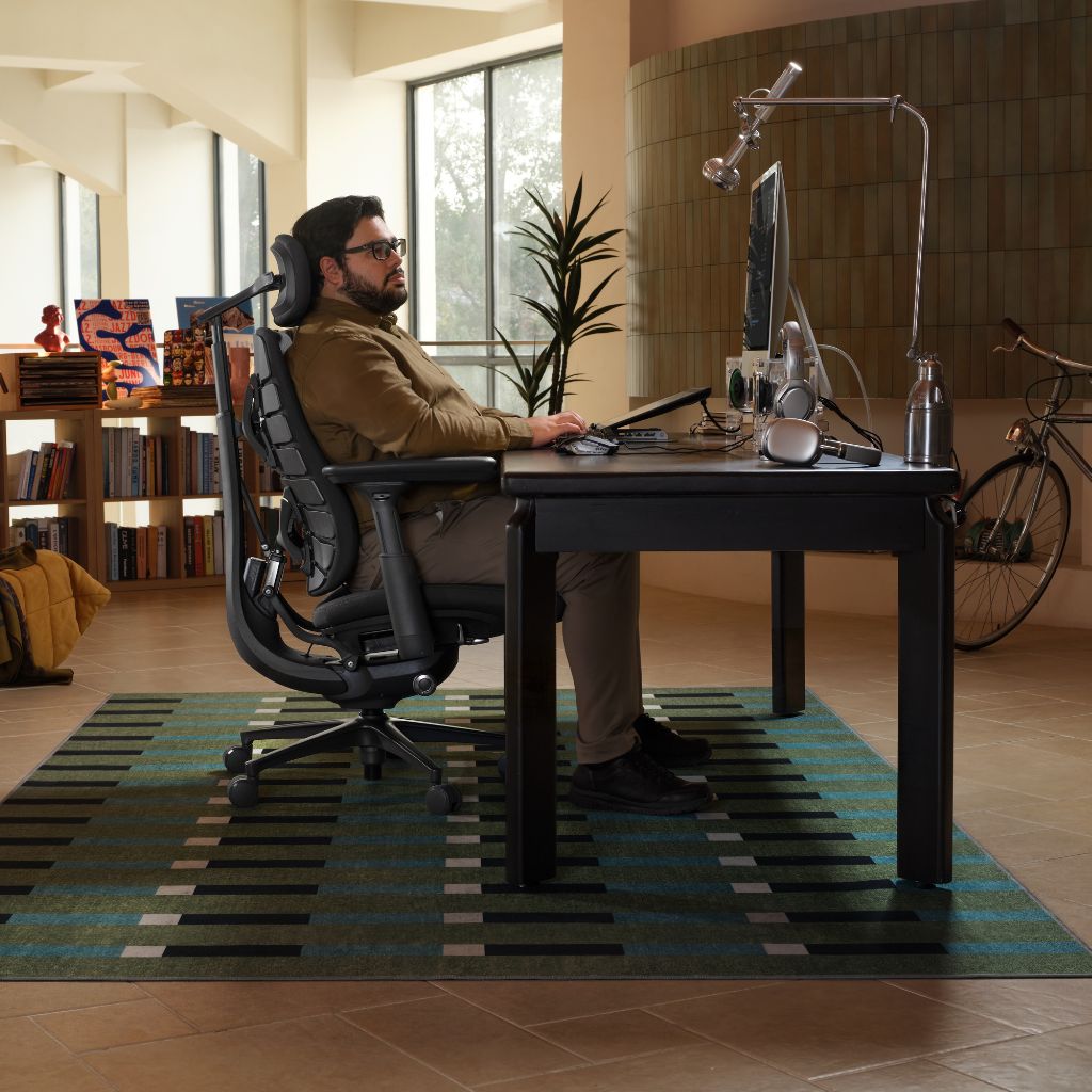 A man sits in a black ergonomic office chair at a dark desk in a modern home office, with a striped rug underneath, a computer monitor, and a bicycle in the background.