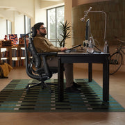 A man sits in a black ergonomic office chair at a dark desk in a modern home office, with a striped rug underneath, a computer monitor, and a bicycle in the background.