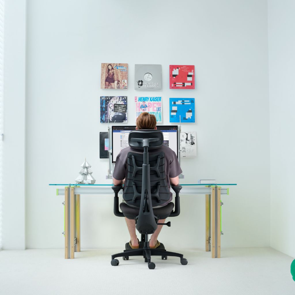 A person sits in a black ergonomic office chair at a glass-top desk, viewed from behind. The room has white walls, a computer monitor on the desk, and several colorful framed prints or books mounted on the wall.