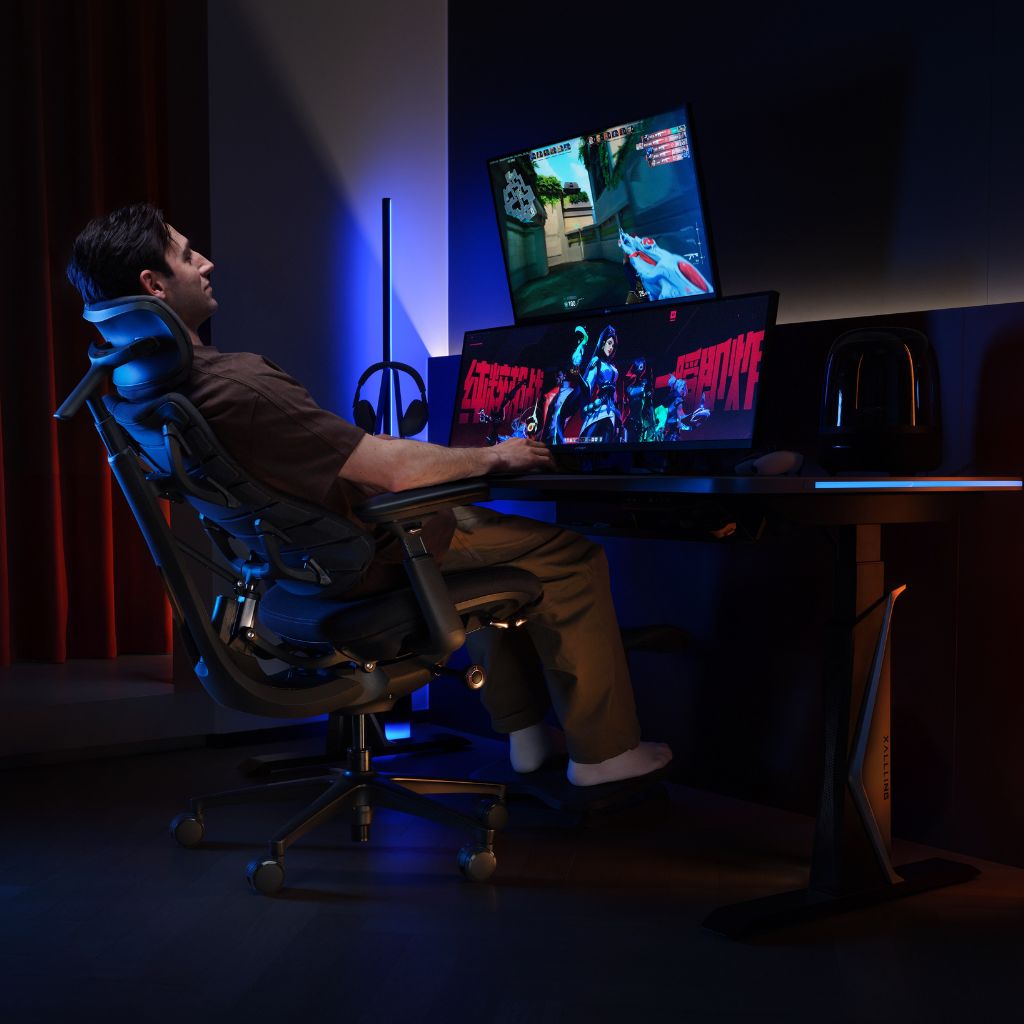 A man sits in a dark gaming chair at a desk with dual computer monitors, illuminated by blue and red ambient lighting in a dim room.