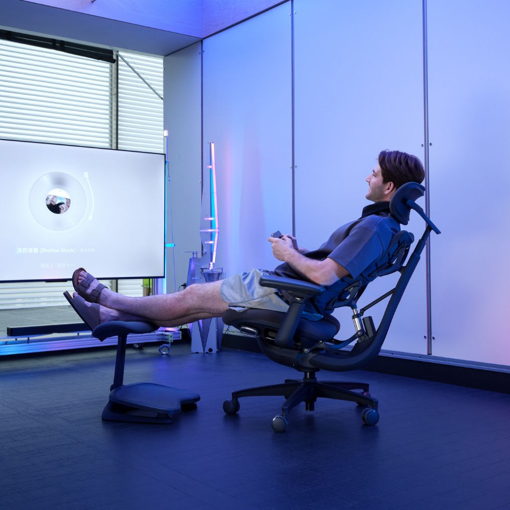 A man reclining in a blue ergonomic office chair with his feet elevated on a matching footrest in a modern room with blue ambient lighting.