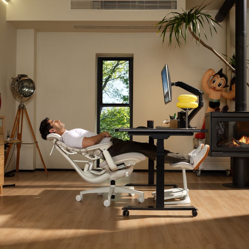 A person reclining in a white ergonomic office chair at a black standing desk with a monitor, in a modern living room with wood floors, a window, and a fireplace.