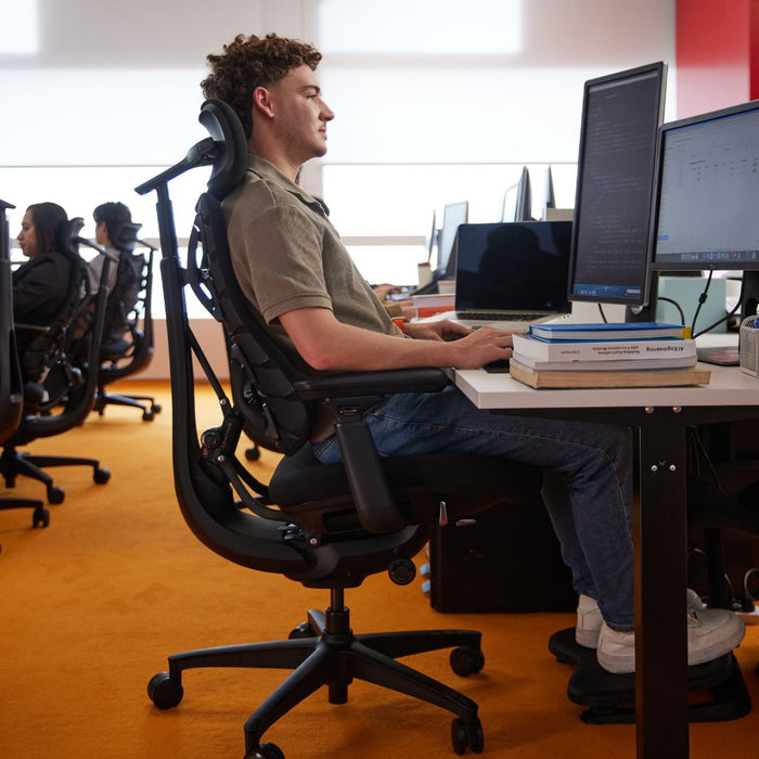 A man sitting in a black ergonomic office chair at a desk with computer monitors, working in a bright office with orange carpet.
