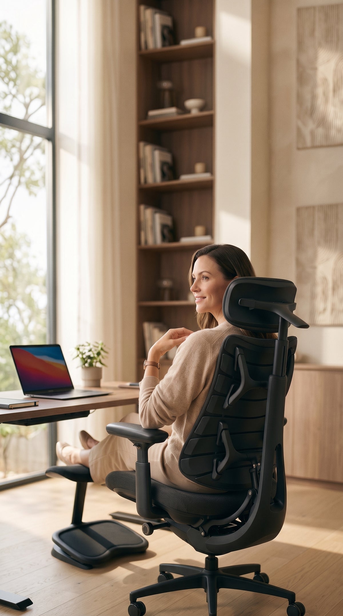 A woman sits in a black ergonomic office chair with a footrest, positioned in a bright, modern home office with wooden bookshelves and a large window.