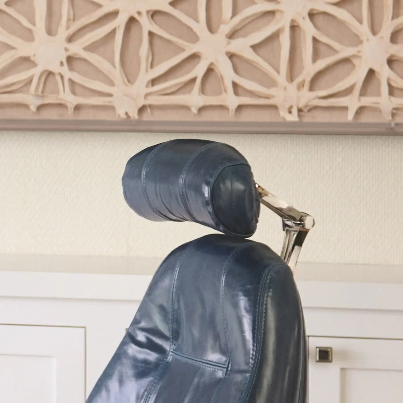 A close-up of a dark blue leather office chair shows a contoured headrest mounted on a polished metal support, set against a textured wall and carved wood artwork in the background.