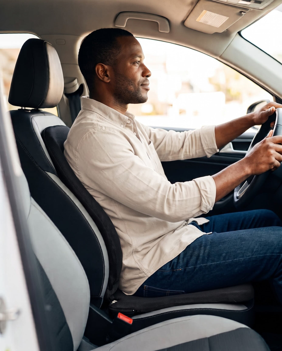 A man driving a car, wearing a light beige long-sleeve shirt and blue jeans, seated in the driver's seat.
