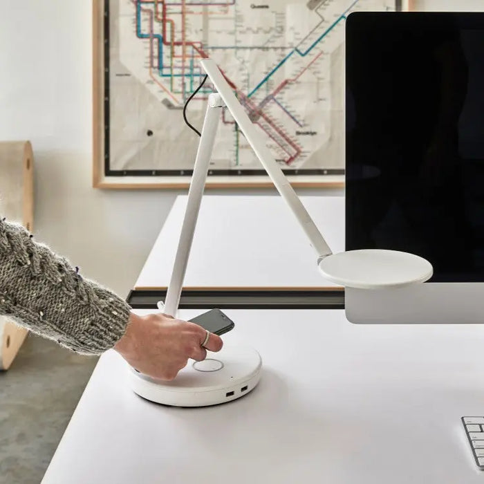 A white adjustable desk lamp with a circular base and USB ports is placed on a white desk next to a monitor, as a person reaches to place a phone on the wireless charging pad.