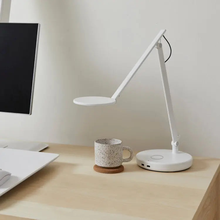 A white adjustable desk lamp with a circular head and base sits on a light wood desk next to a speckled ceramic mug and a computer monitor.