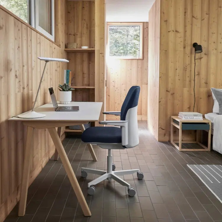 A navy blue and white ergonomic office chair is positioned at a minimalist wooden desk in a cozy, wood-paneled room with natural light and modern decor.