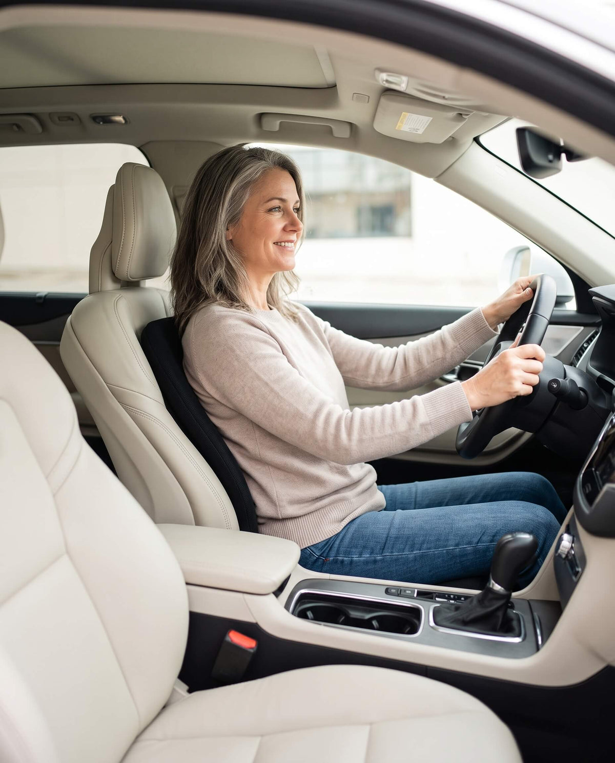 A woman driving a car with cream-colored leather seats, holding the steering wheel and smiling.