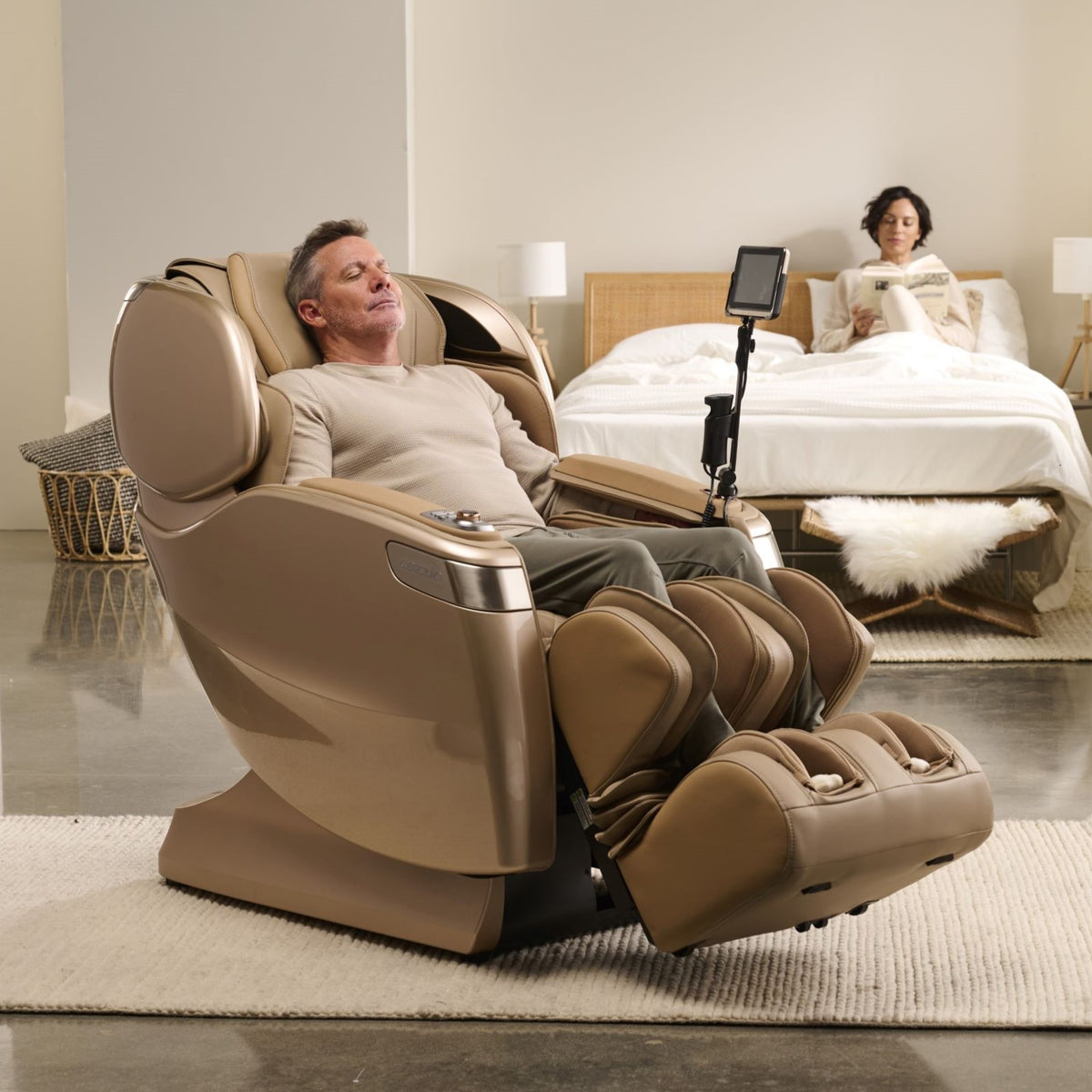 Man relaxing in a massage chair with a woman reading on a bed in the background.