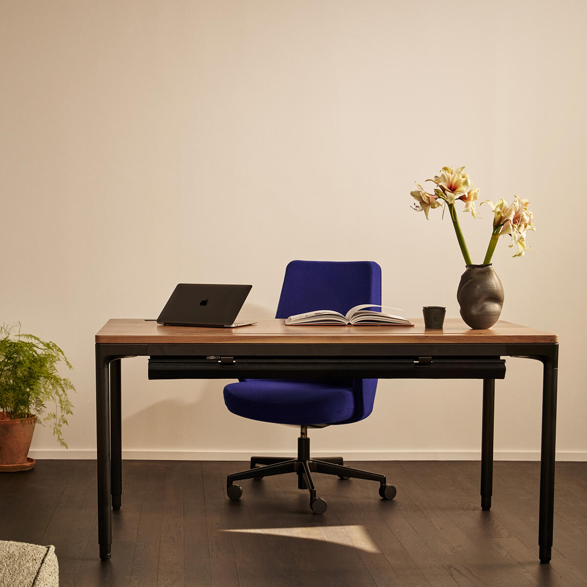 Home office setup with a wooden desk, blue chair, laptop, book, and vase with flowers.