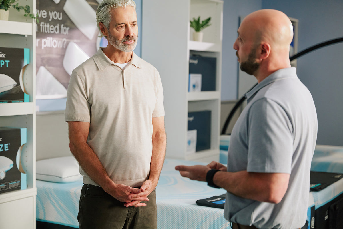 Customer speaking with a sleep specialist beside a mattress display in a Relax The Back store.

