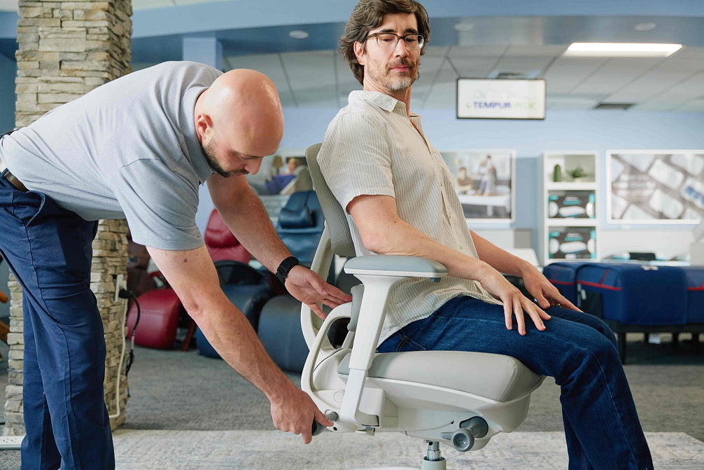A man sits in a light gray ergonomic office chair while another man adjusts the chair's armrests in what appears to be an office showroom or showroom. The background shows other furniture pieces and display frames.