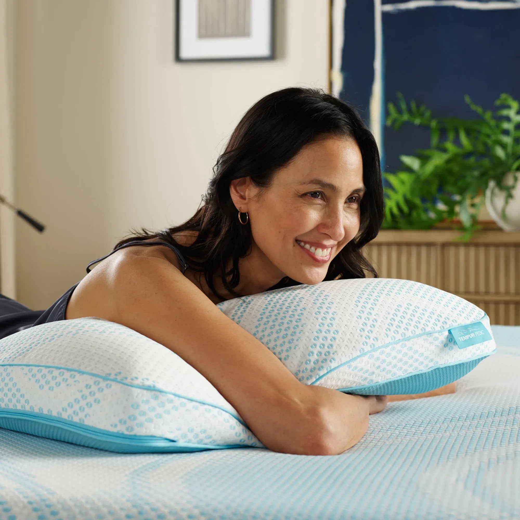 A woman is lying on a bed, smiling while holding the TEMPUR-Breeze® Pro Pillow.