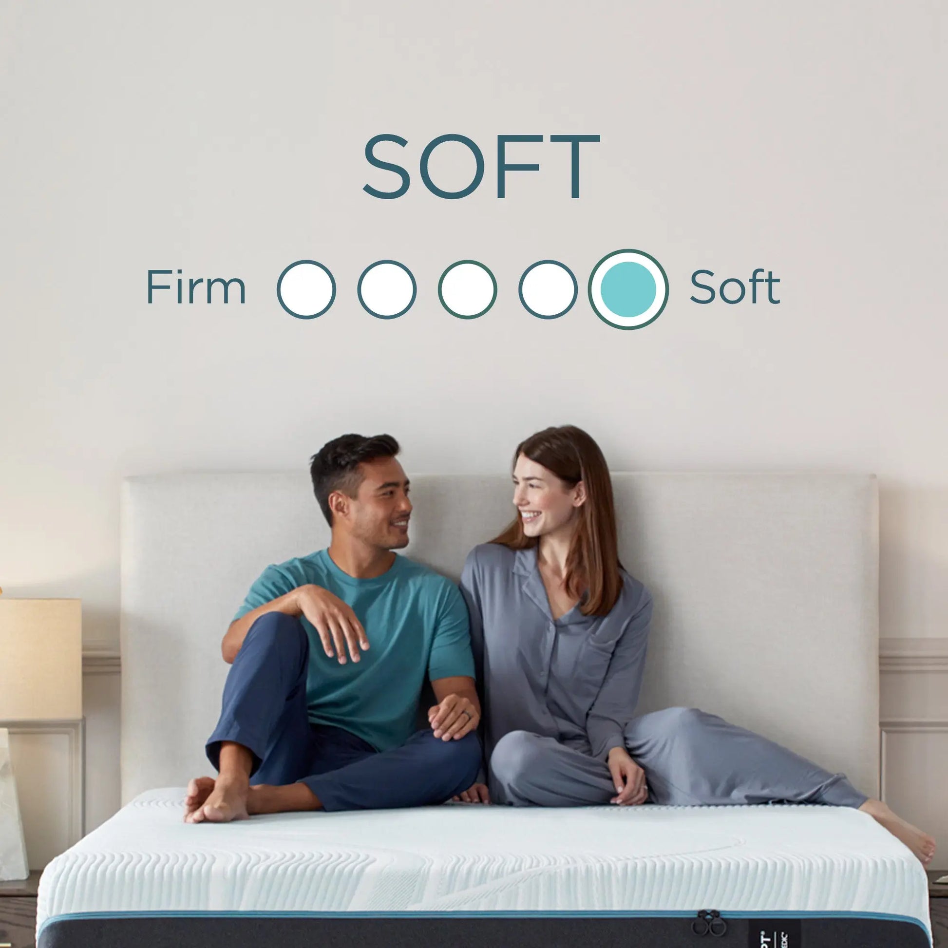 A couple sits on a bed, smiling at each other, with a "Soft" mattress firmness rating displayed above them.