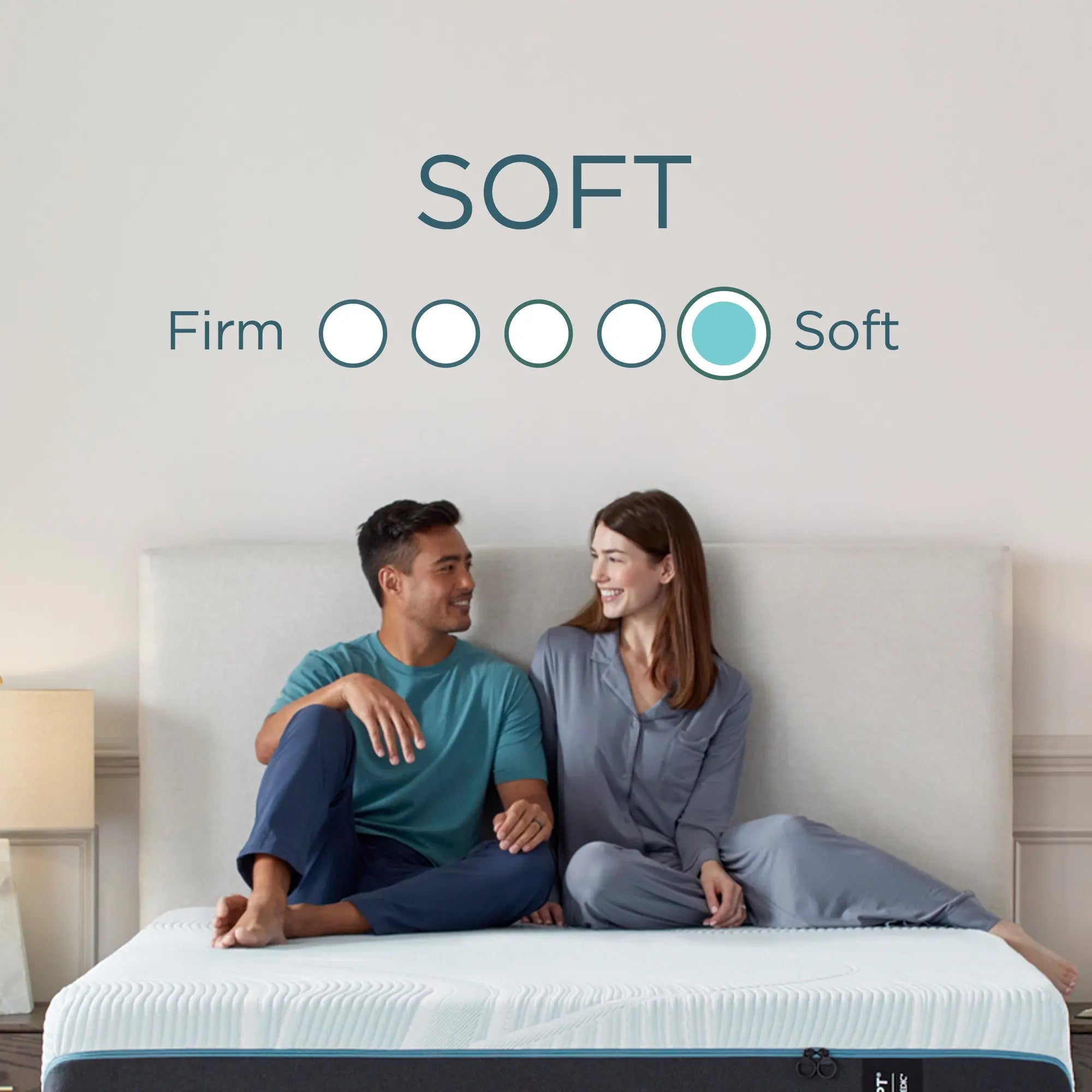 A couple sits on a bed, smiling at each other, with a "Soft" mattress firmness rating displayed above them.