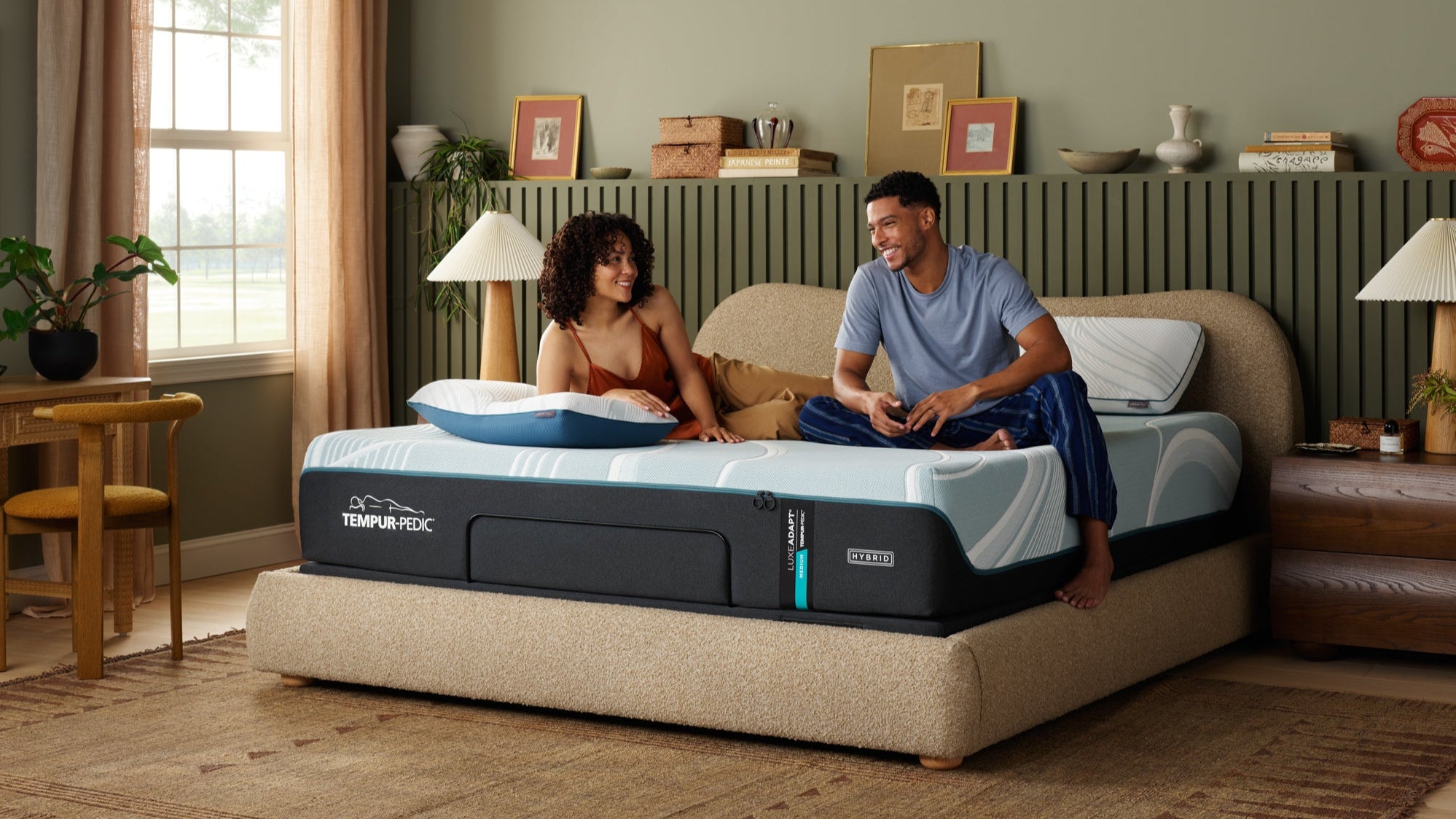 Man and woman sitting on a Tempur-Pedic mattress in a bedroom setting.