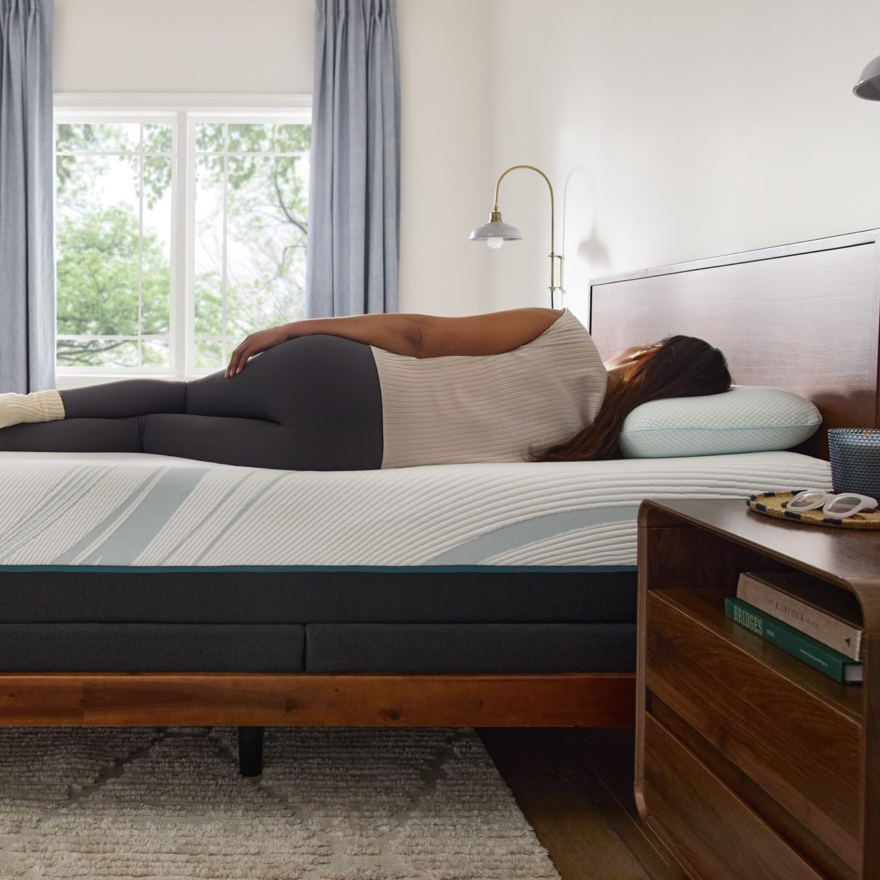 Person lying on a mattress in a bedroom with wooden furniture and large windows.