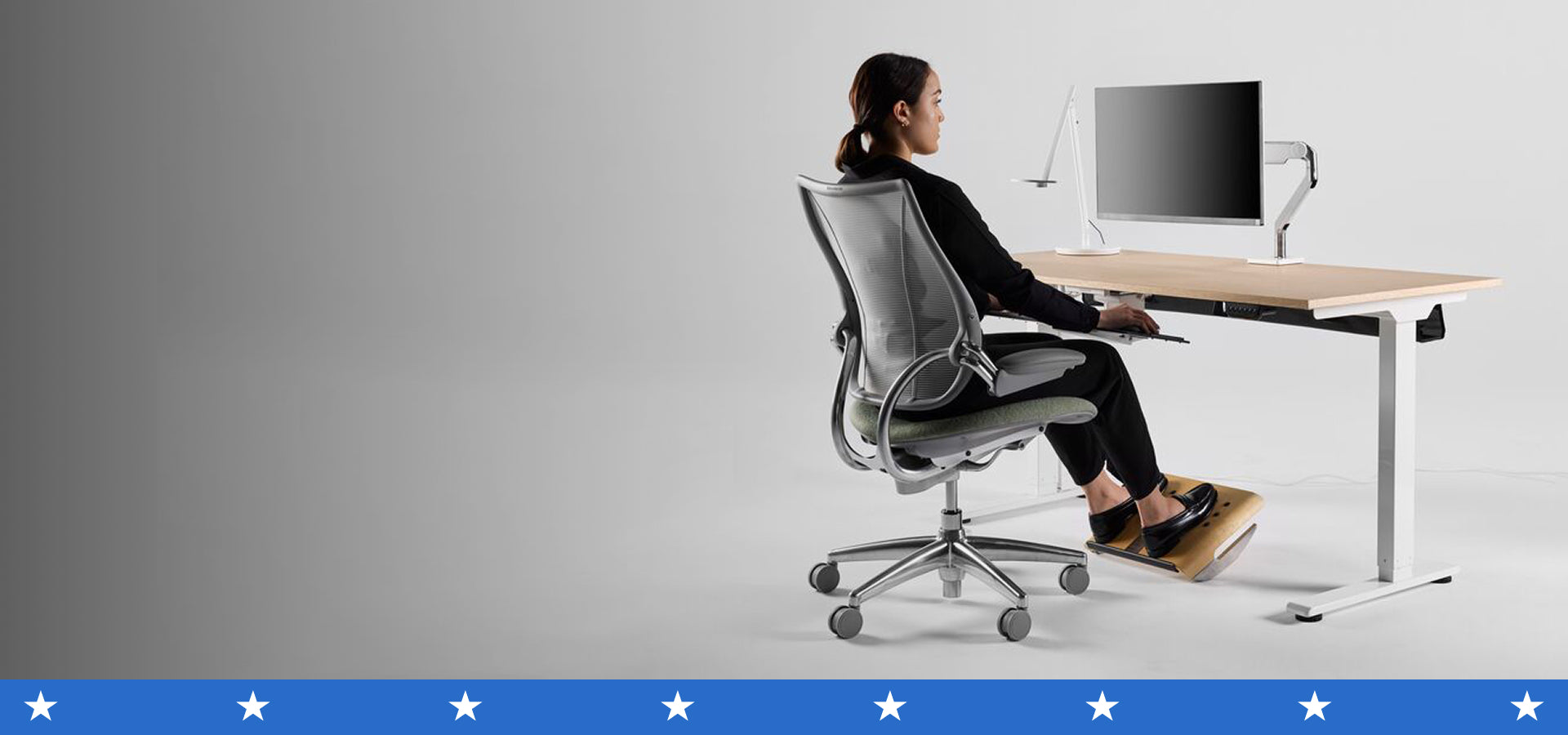 Woman seated ergonomically at a height-adjustable desk with her feet supported by a wooden footrest.