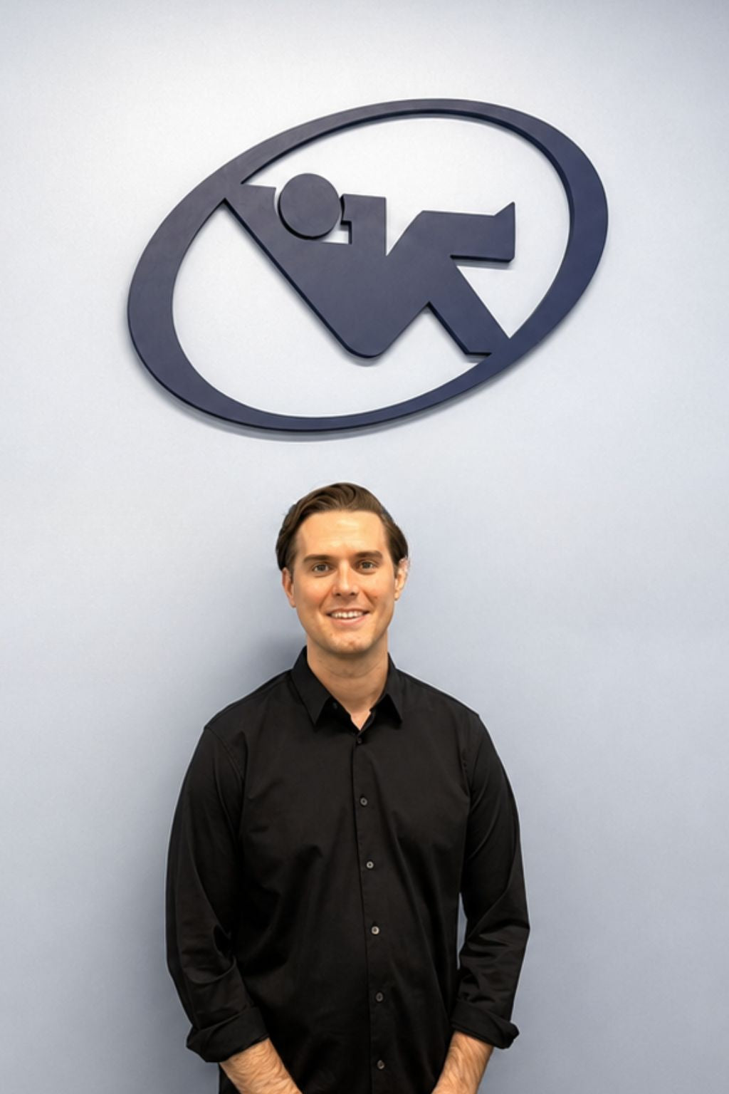 Man standing in front of a wall-mounted Relax The Back logo, wearing a black button-down shirt.